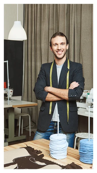 Smiling male tailor standing confidently with arms crossed in a garment workshop, wearing a measuring tape around his neck